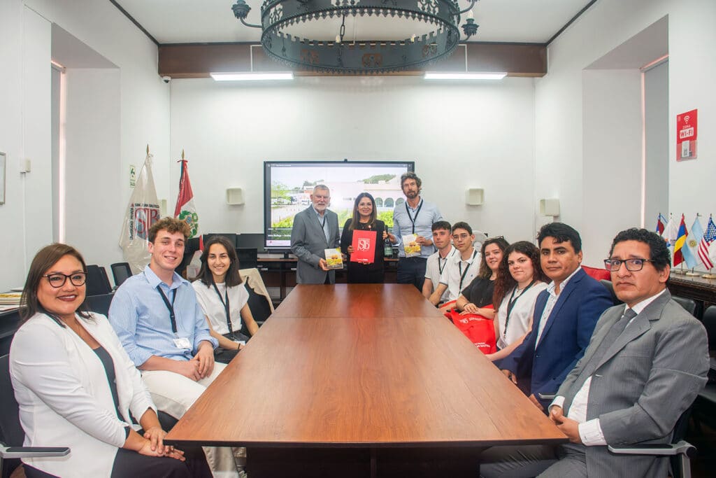 Foto grupal de autoridades de la UPSJB, docentes italianos y estudiantes de ambos países posando alrededor de una gran mesa de madera en una sala de conferencias con una lámpara de araña antigua. Sostienen carpetas y material institucional.