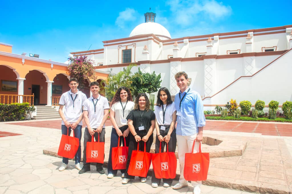 Seis estudiantes italianos posando en un patio colonial español de la UPSJB Campus Hacienda Villa, bajo un cielo azul brillante. Llevan bolsas de regalo rojas con el logo de la UPSJB.