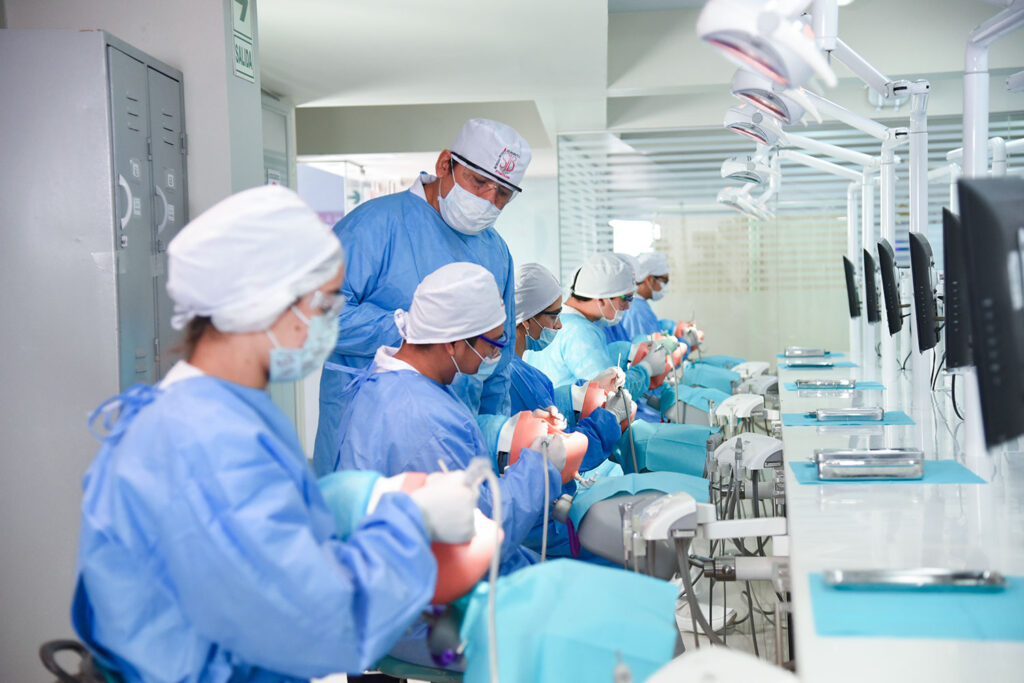 Varios estudiantes de odontología, vestidos con uniformes azules y gorros quirúrgicos blancos, practican en maniquíes dentales alineados en un largo mostrador en un laboratorio espacioso. Un instructor masculino, también con uniforme azul, supervisa a un estudiante. Lámparas dentales cuelgan sobre cada estación de trabajo. El logo "SJB" es visible en algunos gorros.