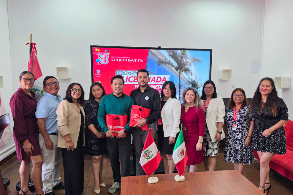 Internacionalización en Tecnología Médica: 5 claves que impulsan la excelencia UPSJB 2 Un grupo de profesionales de la UPSJB y el Mg. Ricardo Salas posan juntos en una sala de reuniones. En primer plano, las banderas de Perú y México. De fondo, una pantalla con información sobre el licenciamiento de la universidad y computadoras de escritorio.