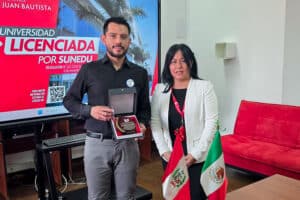 internacionalización en Tecnología Médica UPSJB. La Directora de Tecnología Médica de la UPSJB, Mg. Martha Reyes Coronado, posa junto al ponente internacional de México, Mg. Ricardo Salas, quien sostiene un reconocimiento. De fondo, una pantalla con el logo de la universidad licenciada y una bandera peruana.