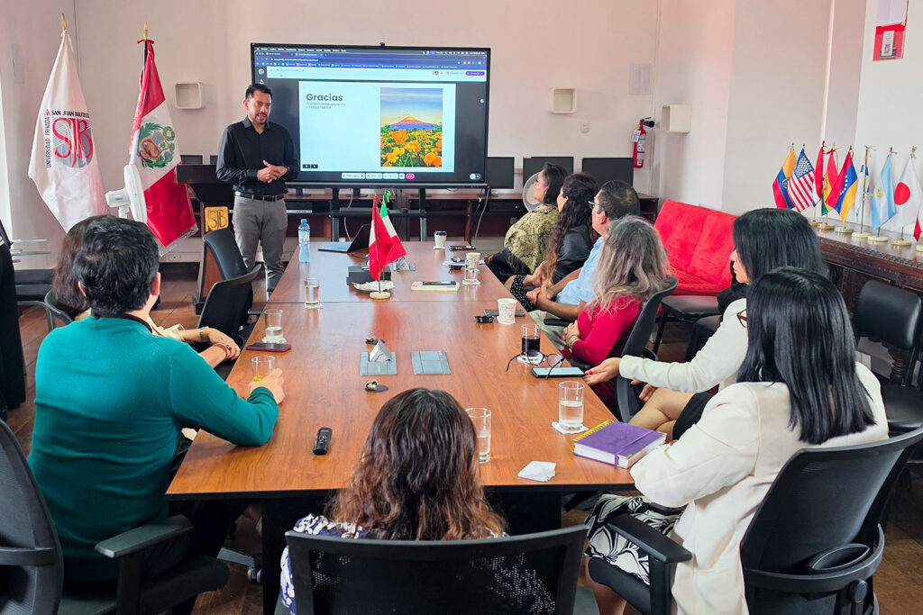 Internacionalización en Tecnología Médica: 5 claves que impulsan la excelencia UPSJB 1 El Mg. Ricardo Salas, de pie frente a una gran pantalla, realiza una presentación ante un grupo de personas sentadas alrededor de una mesa de conferencias en una sala de reuniones. La pantalla muestra una diapositiva con texto y una imagen. Se ven banderas de Perú y otros países en la sala.