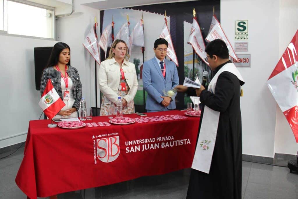 Ceremonia de Ingresantes de Psicología UPSJB: Compromiso y Vocación en Ica y Chincha 2 Una vista de la mesa de honor en un auditorio. Tres autoridades universitarias están de pie detrás de una mesa con mantel rojo y el logotipo de la "UNIVERSIDAD PRIVADA SAN JUAN BAUTISTA". Un sacerdote con sotana negra y estola blanca está de pie a la derecha, dirigiendo la palabra. Banderas peruanas e institucionales están al fondo.