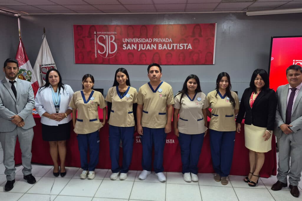Un grupo de estudiantes de Tecnología Médica de la filial Ica de la UPSJB posan para una foto de grupo en una sala de conferencias. Todos llevan sus nuevas batas blancas, con el logo de la universidad. La Mag. Martha Reyes, directora de la filial, y el Dr. [Nombre del director de la escuela], director de la escuela profesional, se encuentran en los extremos de la fila, sonriendo.