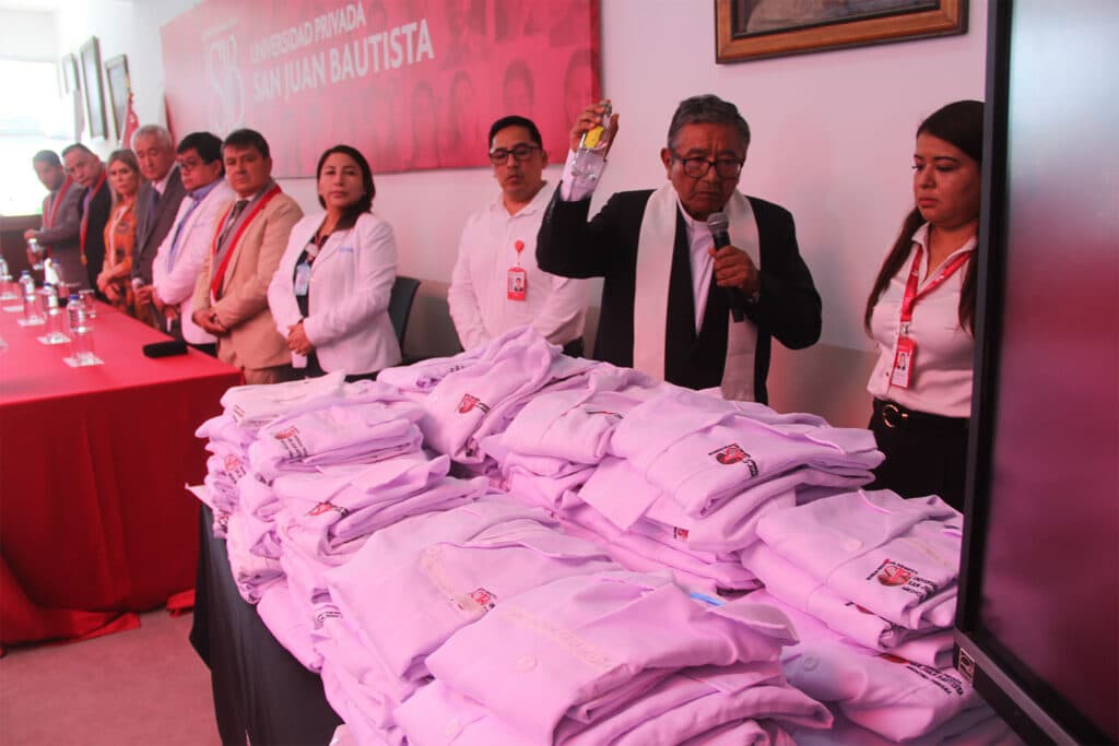 Un grupo de autoridades académicas e invitados de la Escuela Profesional de Medicina Humana de la Universidad Privada San Juan Bautista (UPSJB) se encuentran en fila en la Sala de Grados. Al fondo, un banner con el logo de la universidad y las fotos de los egresados de la promoción. En primer plano, una mesa con las batas blancas de los egresados dobladas y una cruz plateada sobre ellas. Un sacerdote, con su hábito y estola, se encuentra detrás de la mesa, con un micrófono en la mano, y rocía agua bendita sobre las batas.