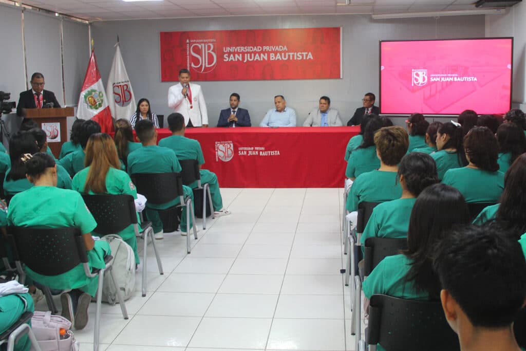Una vista amplia de un auditorio lleno de estudiantes de Medicina Veterinaria de la filial Ica de la UPSJB, vestidos con uniformes médicos verdes y sentados en filas de sillas negras. Al fondo, una mesa presidencial cubierta con un mantel rojo, donde están sentadas varias autoridades académicas e invitados. Un orador en traje blanco habla desde la mesa. Un banner rojo con el logo de la universidad cuelga detrás de la mesa presidencial. Una gran pantalla muestra el mismo logo.