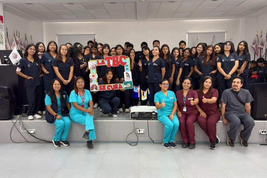 Un grupo numeroso de estudiantes universitarios, la mayoría con uniformes médicos azul marino, posa de pie en un escenario bajo. Al frente, sentados en el borde del escenario, hay cinco profesionales de salud y otro estudiante, sonriendo y con los pulgares arriba. El estudiante sentado en el centro sostiene un marco de campaña de "LA TBC SE CURA". Banderas peruanas y de la UPSJB se ven a los lados.