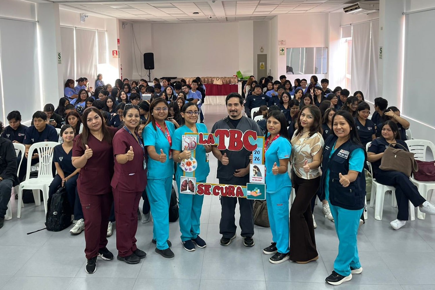 Un grupo de siete profesionales de salud posa en el centro de un auditorio de la UPSJB. Usan uniformes médicos celestes y guindas. En el centro sostienen un marco decorativo que dice "LA TBC SE CURA", "2026", y tiene el logo de la UPSJB. Al fondo, una gran audiencia de estudiantes sentados.