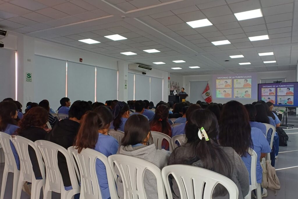 Un grupo numeroso de estudiantes universitarios, la mayoría con uniformes médicos azul marino, posa de pie en un escenario bajo. Al frente, sentados en el borde del escenario, hay cinco profesionales de salud y otro estudiante, sonriendo y con los pulgares arriba. El estudiante sentado en el centro sostiene un marco de campaña de "LA TBC SE CURA". Banderas peruanas y de la UPSJB se ven a los lados.