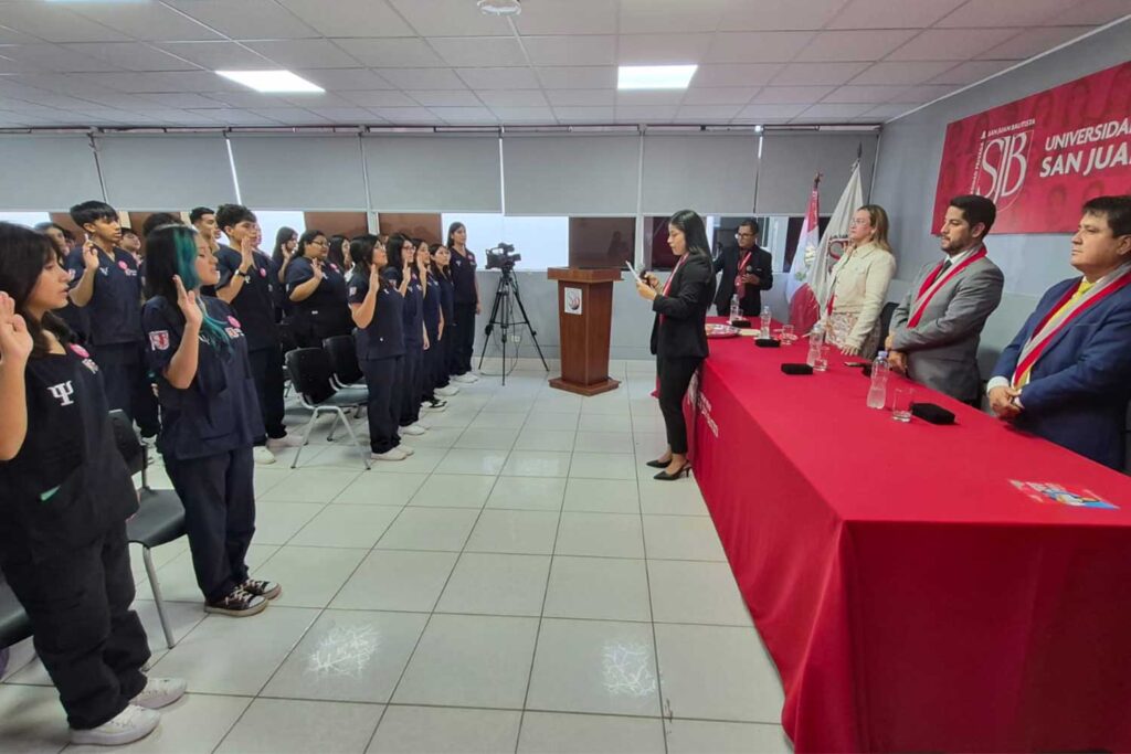 Ceremonia de Ingresantes de Psicología UPSJB: Compromiso y Vocación en Ica y Chincha 4 Vista de un auditorio donde un grupo de estudiantes de Psicología, vestidos con uniformes azul marino, están de pie con la mano derecha levantada en señal de juramento. Frente a ellos, una mesa de honor con mantel rojo donde se encuentran las autoridades universitarias de pie y una coordinadora leyendo el acta de juramentación.