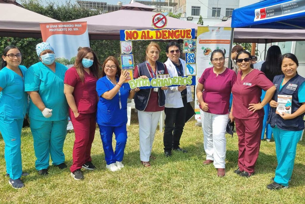 Unidos contra el Dengue: UPSJB impulsa una cultura de prevención y cuidado de la salud en Chorrillos 5 Un grupo de nueve profesionales de la salud posan de pie en un jardín. En el centro, sostienen un marco decorativo con el lema "No al dengue" y un cartel que dice "Evita criaderos". Visten diversos uniformes médicos en colores turquesa, azul y guinda.