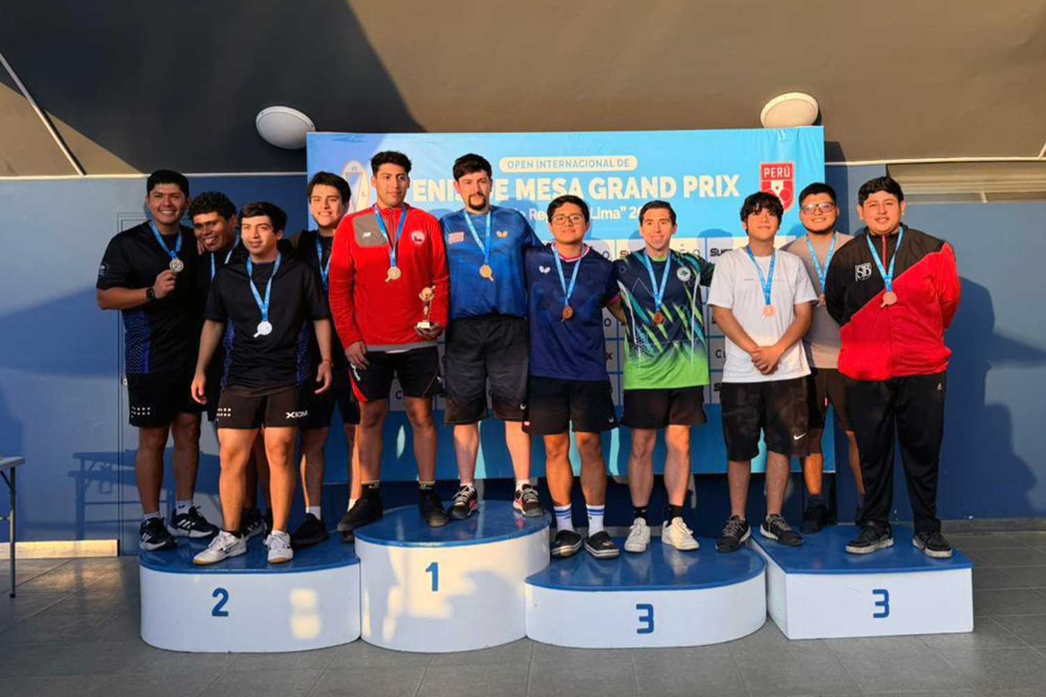 Un grupo de ocho estudiantes hombres de la UPSJB en un podio recibiendo medallas de bronce en el Open Internacional de Tenis de Mesa 2026.
