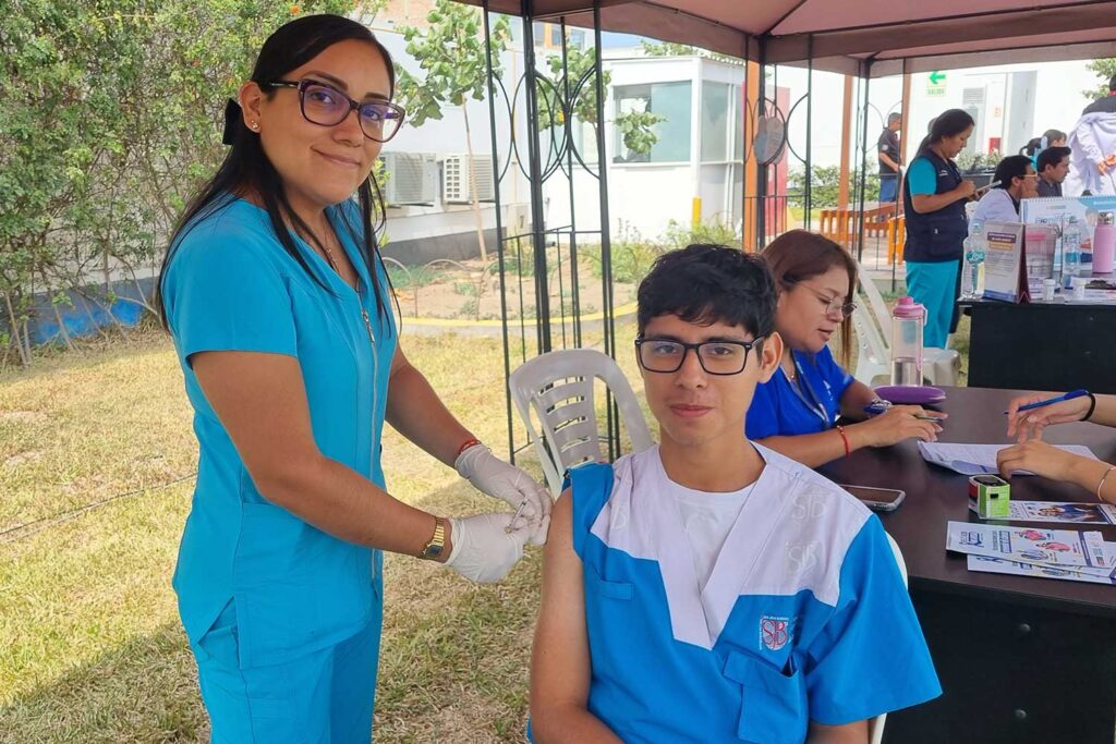 Unidos contra el Dengue: UPSJB impulsa una cultura de prevención y cuidado de la salud en Chorrillos 3 Una enfermera con uniforme turquesa y guantes de protección administra una vacuna en el brazo de un estudiante sentado que viste el uniforme azul y blanco de la UPSJB. Al fondo se observa una mesa con suministros médicos y otras personas en módulos de