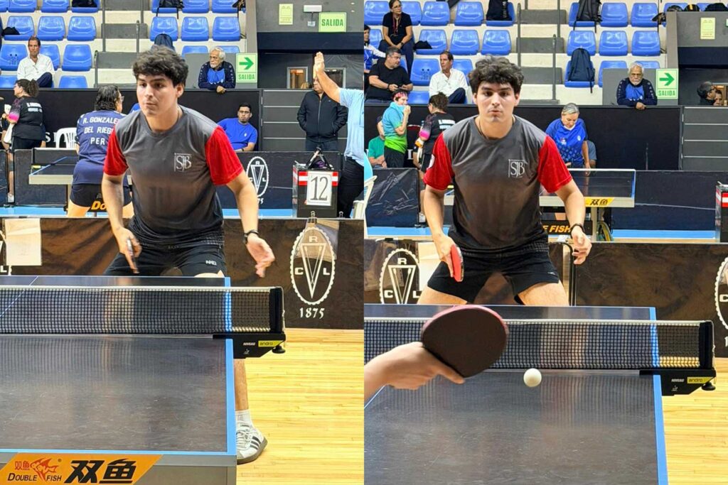 Gianfranco José Chau Izarra, un estudiante de la UPSJB que lleva una camiseta deportiva gris y roja con el logo de la universidad, está en una pose dinámica, a punto de golpear una pelota de tenis de mesa sobre la red con su paleta. La pelota está en el aire. El fondo está ligeramente borroso, mostrando la sala de tenis de mesa con otras mesas y personas. El mismo cartel azul con el logo de la UPSJB es visible en la distancia.