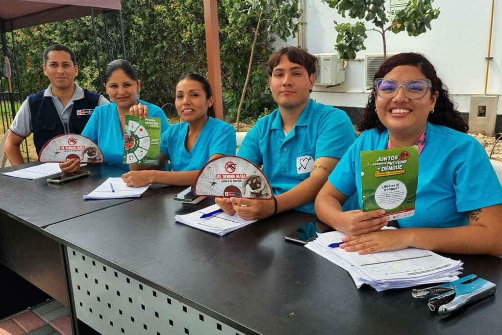 Unidos contra el Dengue: UPSJB impulsa una cultura de prevención y cuidado de la salud en Chorrillos 1 Cinco personas sentadas tras un escritorio en un espacio exterior. Cuatro de ellas visten uniformes médicos turquesa y muestran folletos preventivos con mensajes como "Juntos podemos prevenir el dengue" e "¡El dengue mata!". Sobre la mesa hay lapiceros, hojas de registro y una engrapadora.