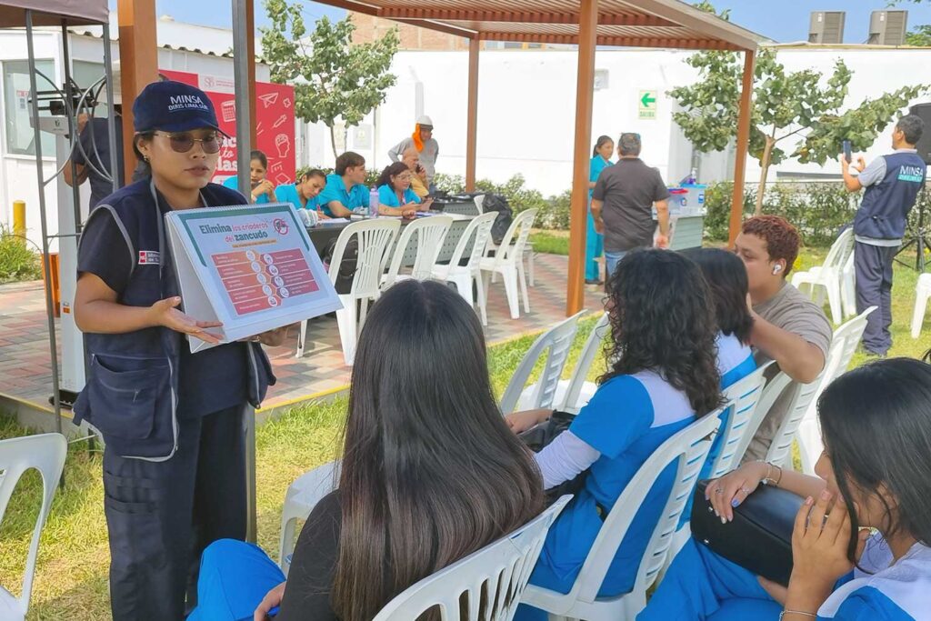 Unidos contra el Dengue: UPSJB impulsa una cultura de prevención y cuidado de la salud en Chorrillos 2 Una facilitadora de salud con gorra y chaleco del MINSA explica un rotafolio titulado "Elimina los criaderos del zancudo" ante un grupo de estudiantes sentados. La actividad se realiza al aire libre bajo una estructura de madera.