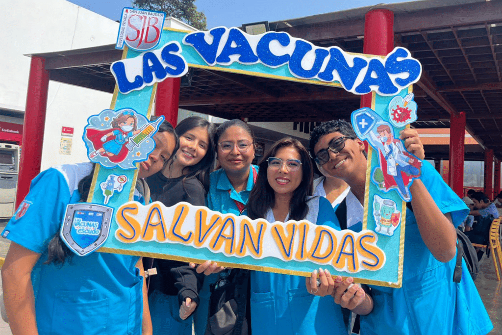 Unidos contra el Dengue: UPSJB impulsa una cultura de prevención y cuidado de la salud en Chorrillos 4 Seis jóvenes sonrientes posan detrás de un marco colorido y brillante que dice "Las vacunas salvan vidas". El marco incluye ilustraciones de personal de salud como superhéroes y el escudo de la Universidad Privada San Juan Bautista (UPSJB).