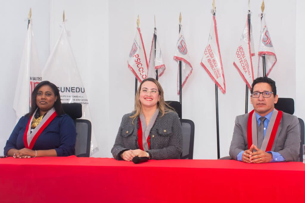 autoridades-academicas-escuela-psicologia-upsjb Toma de plano medio que muestra a las tres principales autoridades del evento sentadas detrás de una mesa con mantel rojo. Portan las medallas académicas con cintas rojas de la Universidad Privada San Juan Bautista. Al fondo se aprecian estandartes blancos impresos con las distintas facultades y escuelas, incluyendo Psicología y SUNEDU.