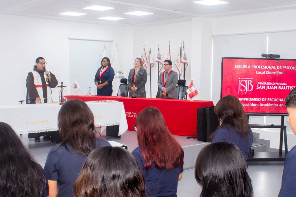 paraliturgia-investidura-academica-psicologia-chorrillos Vista desde atrás de los estudiantes presenciando la paraliturgia oficiada por un sacerdote frente a las autoridades académicas en la mesa de honor de la Ceremonia de Investidura Académica en Psicología.