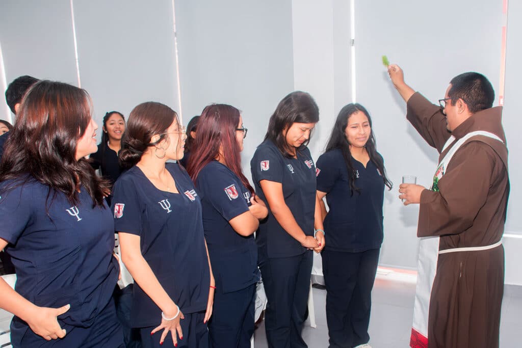 bendicion-de-chaquetas-pines-psicologia-upsjb Sacerdote Víctor Hugo Conce Yauri rociando agua bendita sobre los uniformes de las estudiantes de psicología de la UPSJB.