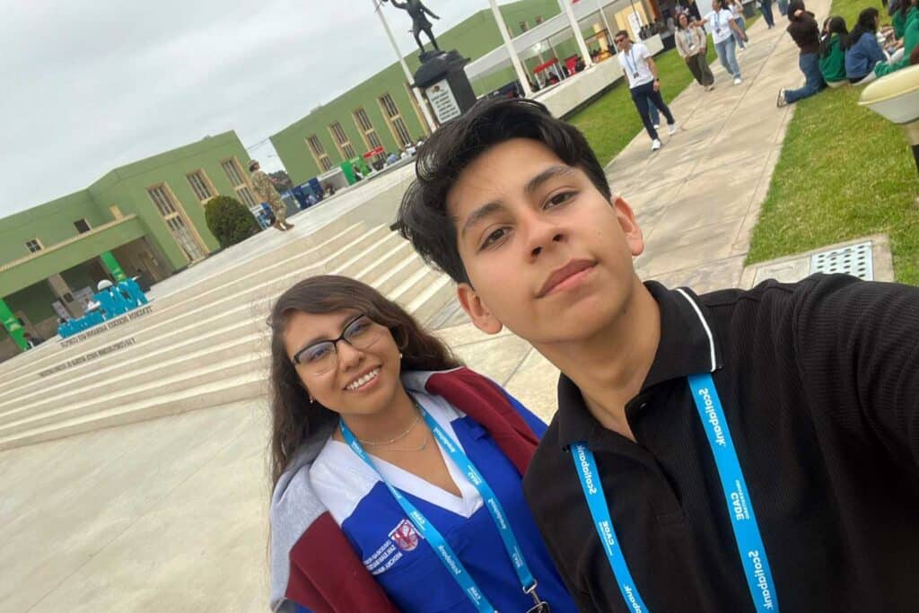 Una selfie de Jesús Descalzo y Iris Tasayco, estudiantes de la UPSJB, en la Escuela Militar de Chorrillos. Ambos sonríen a la cámara. Jesús, en primer plano a la derecha, usa una polo negra. Iris, a la izquierda, tiene lentes y una chaqueta. De fondo, se ve un edificio verde, escaleras de concreto y una estatua en lo alto, bajo un cielo nublado.