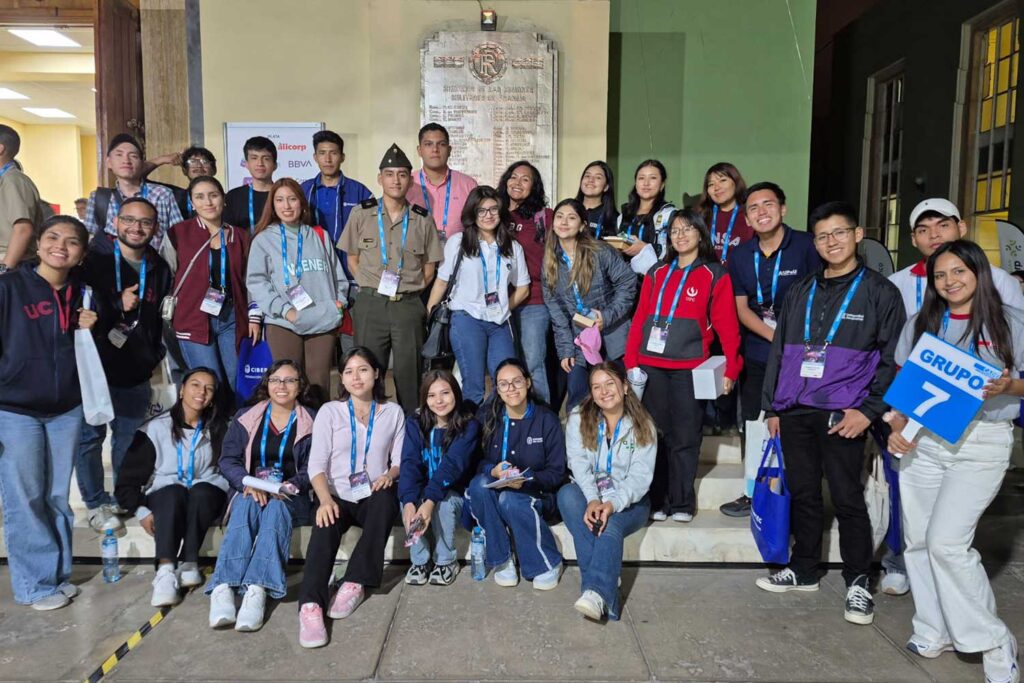 Una foto grupal nocturna con más de 20 estudiantes posando en unas escaleras de concreto frente a un edificio con una placa conmemorativa. Iris Tasayco está sentada en primera fila, con lentes y chaqueta oscura. Un estudiante a la derecha sostiene un cartel azul que dice "GRUPO 7 CADE". Varios estudiantes llevan credenciales y bolsas de tela azules.