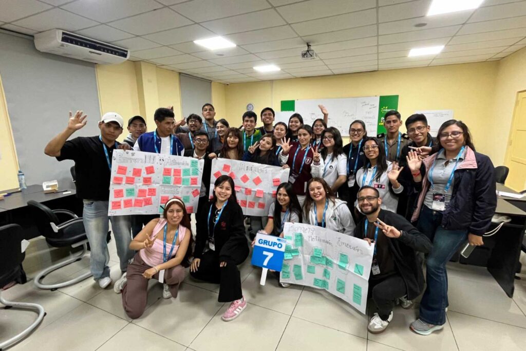 Un grupo de más de 20 estudiantes posa en un aula bien iluminada. En el centro, muestran tres grandes carteles de papel llenos de notas adhesivas rojas y verdes, titulados "Barreras..." y "Soluciones...". Iris Tasayco está de pie a la derecha, sonriendo. Un estudiante sostiene un cartel que dice "GRUPO 7 CADE". Otros hacen señas de paz o saludan.