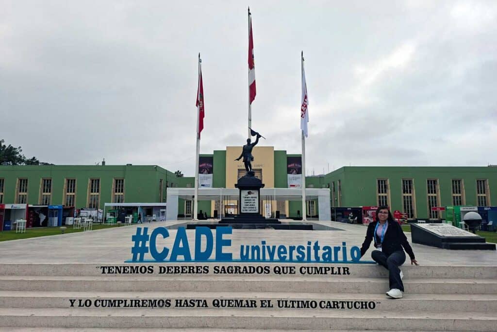 Una foto de Iris Tasayco sentada en las escaleras frente a un gran edificio verde con tres banderas (Perú y otras dos blancas). Ella sonríe y tiene las piernas cruzadas. En las escaleras se leen letras azules grandes: "#CADE universitario" y debajo, en negro: "TENEMOS DEBERES SAGRADOS QUE CUMPLIR / Y LO CUMPLIREMOS HASTA QUEMAR EL ÚLTIMO CARTUCHO".