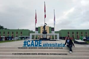Una foto de Iris Tasayco sentada en las escaleras frente a un gran edificio verde con tres banderas (Perú y otras dos blancas). Ella sonríe y tiene las piernas cruzadas. En las escaleras se leen letras azules grandes: "#CADE universitario" y debajo, en negro: "TENEMOS DEBERES SAGRADOS QUE CUMPLIR / Y LO CUMPLIREMOS HASTA QUEMAR EL ÚLTIMO CARTUCHO".
