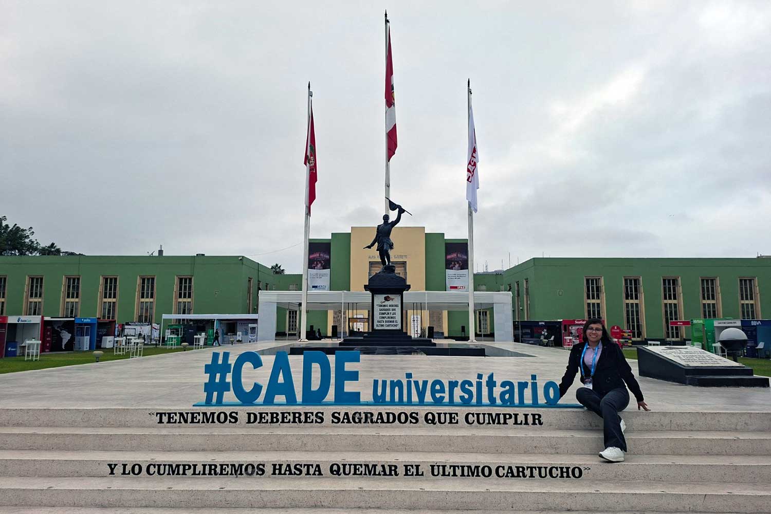 Una foto de Iris Tasayco sentada en las escaleras frente a un gran edificio verde con tres banderas (Perú y otras dos blancas). Ella sonríe y tiene las piernas cruzadas. En las escaleras se leen letras azules grandes: "#CADE universitario" y debajo, en negro: "TENEMOS DEBERES SAGRADOS QUE CUMPLIR / Y LO CUMPLIREMOS HASTA QUEMAR EL ÚLTIMO CARTUCHO".