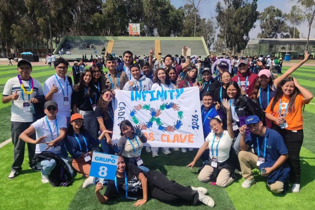 Una foto grupal de unos 25 estudiantes posando en un campo de césped artificial bajo la luz del sol. Varios sostienen una gran pancarta blanca que dice "INFINITY" y "CADE 2026" con huellas de manos de colores. Jesús Descalzo está de pie detrás de la pancarta. Un estudiante en el suelo sostiene un cartel que dice "GRUPO 8 CADE".
