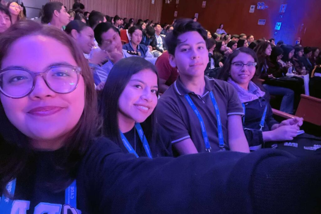 Una selfie grupal tomada por una estudiante en un auditorio oscuro con iluminación azul y morada. Ella está en primer plano a la izquierda. Detrás de ella, a la derecha, Jesús Descalzo sonríe, y más atrás, Iris Tasayco también sonríe con sus lentes. Hay una multitud de otros estudiantes sentados en el fondo, desenfocados.