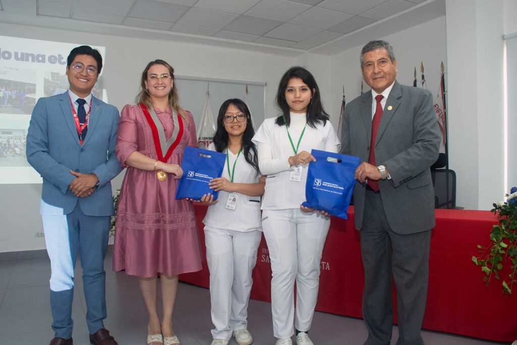 Una fotografía de una ceremonia formal de premiación en interiores. Dos autoridades de la UPSJB, un hombre con un traje azul claro a la izquierda y otro con un traje gris a la derecha, posan con dos estudiantes de enfermería vestidas con uniformes médicos blancos. Las estudiantes sostienen bolsas de regalo azules con el logotipo de la UPSJB. La autoridad femenina central, con un vestido rosa y un collar ceremonial, sonríe a la cámara. Un cartel en la pared de la izquierda dice "o una et". Una mesa con un mantel rojo y arreglos florales está a la derecha.