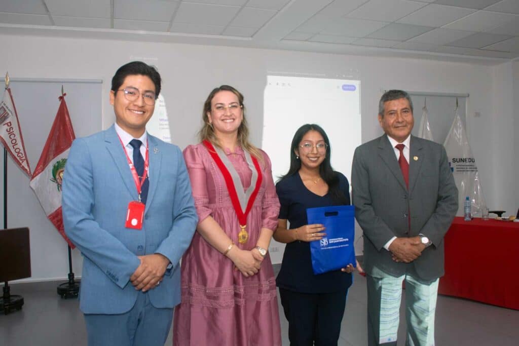 Una fotografía de una ceremonia formal de premiación en interiores. Un grupo de siete personas posa para la cámara. Cinco estudiantes de la UPSJB sostienen certificados y trofeos. Las dos autoridades universitarias, el hombre con traje gris y la mujer con vestido rosa y collar ceremonial, posan con ellos. Un cartel en la pared de la izquierda dice "PSICOLOGÍA". Una mesa con un mantel rojo y arreglos florales está a la derecha. El fondo es una pared blanca con el logotipo de la UPSJB.