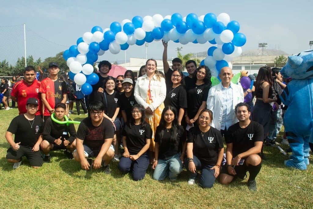 Estudiantes y docentes de Psicología de la UPSJB posando bajo un arco de globos azules y blancos en un campo abierto durante la semana de aniversario.