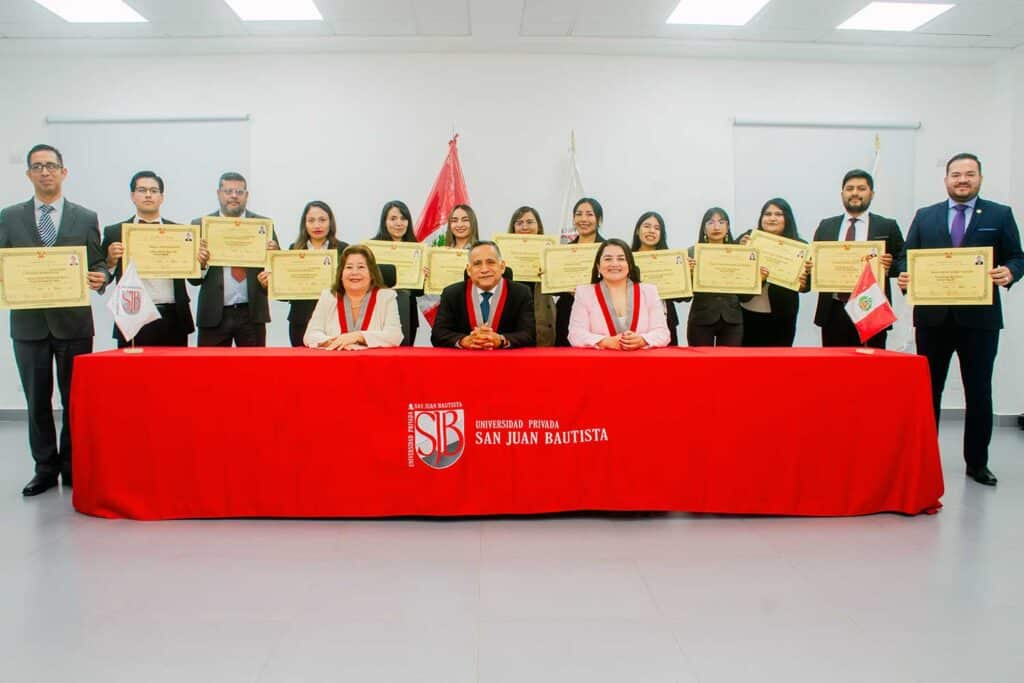 Foto grupal de autoridades de la UPSJB sentadas y un grupo de egresados de posgrado de pie mostrando sus diplomas de maestro y especialista detrás de la mesa de honor.