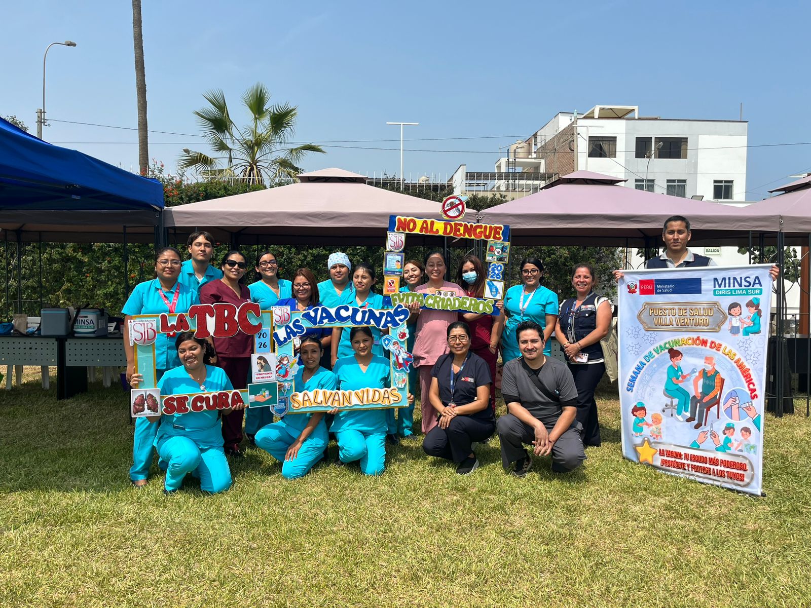 Foto grupal de 15 personas, entre profesionales de salud y personal administrativo, posando en un área verde frente a carpas de atención. Sostienen marcos informativos sobre vacunas y prevención del dengue. A la derecha, un banner del MINSA menciona al Puesto de Salud Villa Venturo.