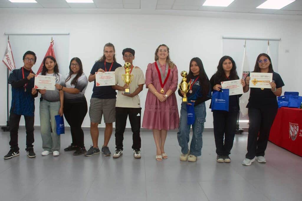 Una fotografía de una ceremonia formal de premiación en interiores. Las mismas cuatro personas de la imagen anterior se colocan en una posición ligeramente diferente. El estudiante de la izquierda con traje azul claro ahora mira a la cámara. La estudiante de enfermería de la izquierda sostiene una bolsa de regalo azul con el logotipo de la UPSJB. La estudiante de la derecha, también con uniforme médico blanco, sonríe a la cámara. Un cartel en la pared de la izquierda dice "UPSJB" y "PSICOLOGÍA". Un cartel a la derecha dice "SUNEDU". Una mesa con un mantel rojo y arreglos florales está a la derecha.