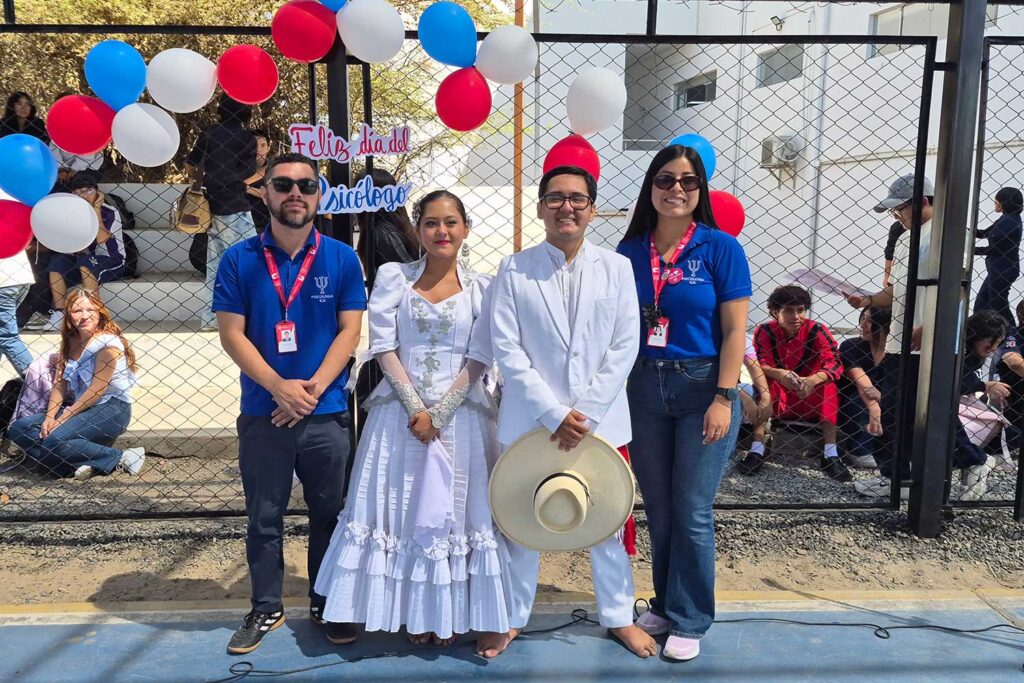 Dos estudiantes vestidos con trajes típicos de marinera (blanco) posan junto a dos docentes con uniformes azules de la UPSJB. Al fondo se ve un cartel que dice "Feliz Día del Psicólogo" y globos decorativos.