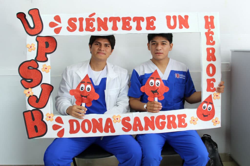 Estudiantes se suman a la sensibilización sobre la donación de sangre Dos estudiantes de ciencias de la salud sostienen un marco decorativo con mensajes de apoyo a la donación de sangre durante una jornada de sensibilización en la universidad.