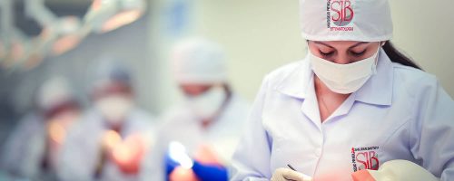 Una estudiante de odontología con mascarilla y guantes trabaja en un maniquí de práctica en un laboratorio clínico de odontología. Otros estudiantes están en el fondo, desenfocados, realizando actividades similares bajo luces clínicas brillantes. El gorro de la estudiante muestra el logo "SJB ESTOMATOLOGIA".
