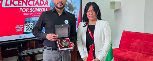 internacionalización en Tecnología Médica UPSJB. La Directora de Tecnología Médica de la UPSJB, Mg. Martha Reyes Coronado, posa junto al ponente internacional de México, Mg. Ricardo Salas, quien sostiene un reconocimiento. De fondo, una pantalla con el logo de la universidad licenciada y una bandera peruana.