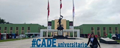 Una foto de Iris Tasayco sentada en las escaleras frente a un gran edificio verde con tres banderas (Perú y otras dos blancas). Ella sonríe y tiene las piernas cruzadas. En las escaleras se leen letras azules grandes: "#CADE universitario" y debajo, en negro: "TENEMOS DEBERES SAGRADOS QUE CUMPLIR / Y LO CUMPLIREMOS HASTA QUEMAR EL ÚLTIMO CARTUCHO".