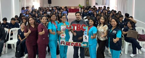 Un grupo de siete profesionales de salud posa en el centro de un auditorio de la UPSJB. Usan uniformes médicos celestes y guindas. En el centro sostienen un marco decorativo que dice "LA TBC SE CURA", "2026", y tiene el logo de la UPSJB. Al fondo, una gran audiencia de estudiantes sentados.
