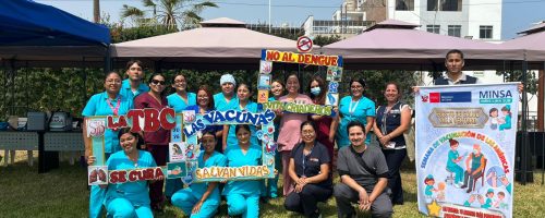 Foto grupal de 15 personas, entre profesionales de salud y personal administrativo, posando en un área verde frente a carpas de atención. Sostienen marcos informativos sobre vacunas y prevención del dengue. A la derecha, un banner del MINSA menciona al Puesto de Salud Villa Venturo.