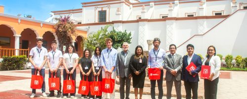 Foto grupal completa de la delegación italiana y autoridades de la UPSJB posando en el patio central del Campus Hacienda Villa, frente al edificio colonial blanco con cúpula y arcos naranjas.